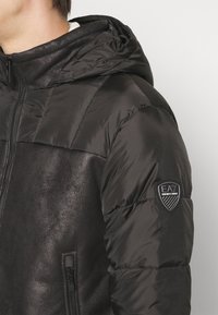 Svart puffjacka med slät yta, utrustad med huva, fickor med dragkedja och en logotypmärke på vänster ärm.