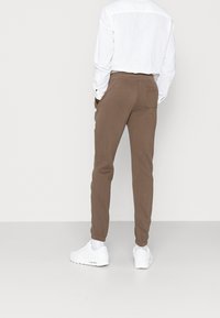 Hnědé jogger kalhoty s úzkým střihem, elastickým pasem a zadní kapsou, v kombinaci s bílou košilí s knoflíky a bílými teniskami.