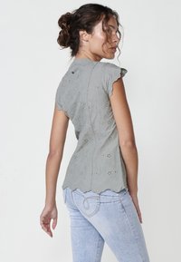 Grau gestickte Bluse mit gewellten Ärmeln und Saum, die florale Muster zeigt; kombiniert mit hellblauen Jeans. Die Textur erscheint weich und detailreich.