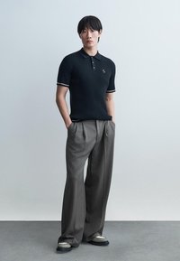 Jeune homme debout les mains dans les poches, portant un polo noir à manches courtes côtelé et un large pantalon gris, sur fond uni.