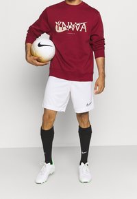 Personne tenant un ballon de football Nike blanc et or, portant un sweat-shirt rouge du Liverpool FC, un short Nike blanc, des chaussettes noires et des crampons blancs.