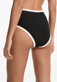 Magas derekú fekete bikini alsó, krémszínű kontraszt díszítéssel, puha anyaggal és sima textúrával, hátulról és oldalról látható.
