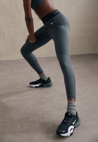 Osoba w szarych, wzorzystych legginsach Nike Pro, szarych skarpetkach i czarnych butach Nike rozciąga się w pomieszczeniu na beżowej podłodze.