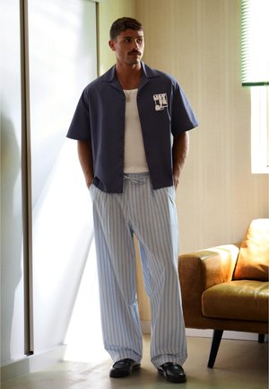 Homme avec moustache debout à l'intérieur portant une chemise bleu marine à manches courtes, un débardeur blanc, un pantalon large rayé bleu et des chaussures noires près d'un fauteuil marron.