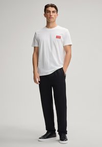 Biała bawełniana koszulka z małym czerwonym kwadratowym logo na lewej piersi, w połączeniu z czarnymi spodniami dresowymi i czarnymi sneakersami. Minimalistyczny design.