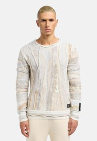 Lys strikket sweater med indviklede mønstre i beige og crème, med ribbede manchetter og kant. Tekstiler med lagdelte designelementer.