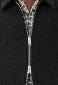Zwarte fleecvest met ritssluiting en een gestructureerd oppervlak, zilveren rits, gedragen over een groen-wit geruit shirt.