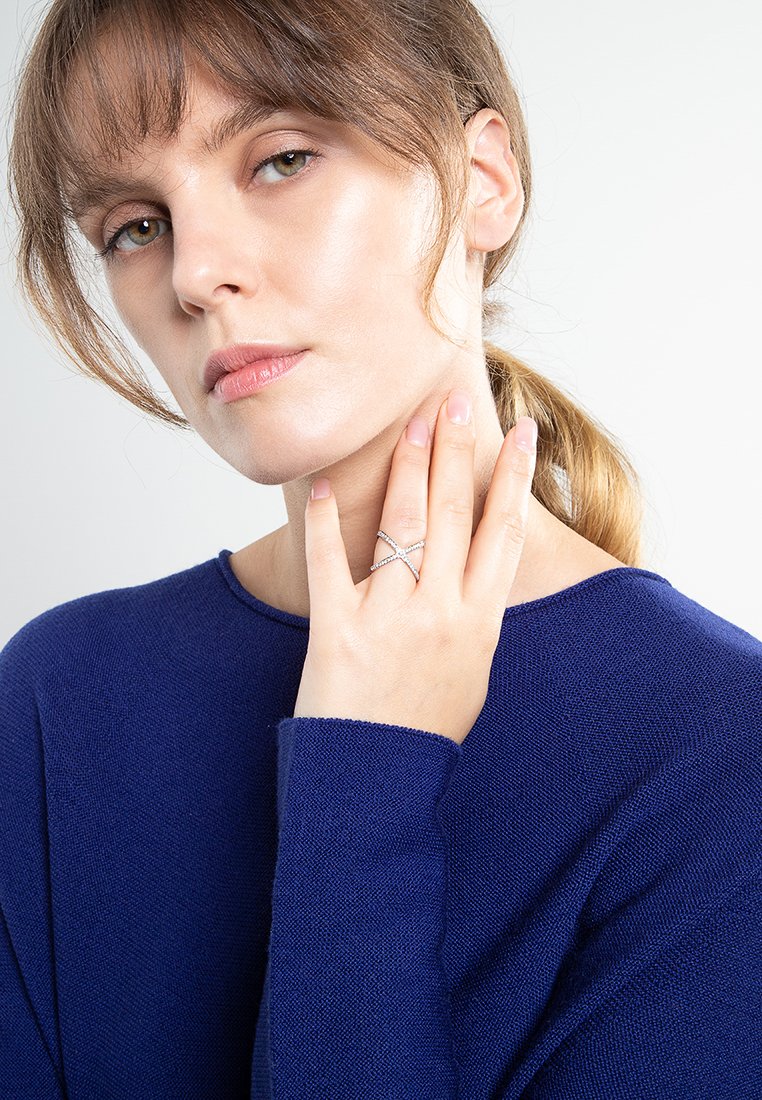 Anillo cruzado de plata con piedras de strass llevado en una mano. El modelo lleva un suéter de punto texturizado en azul marino, con un fondo neutro.