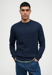 Granatowy sweatshirt z długimi rękawami i okrągłym dekoltem, o gładkiej fakturze. Nosi się go na zielonej koszuli, zestawiony z dżinsowymi spodniami.