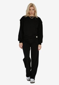 Czarna bluza z luźnym krojem, z prążkowanymi mankietami i małą metką z logo. Zestawiona z pasującymi czarnymi spodniami o szerokich nogawkach i białymi sneakersami.