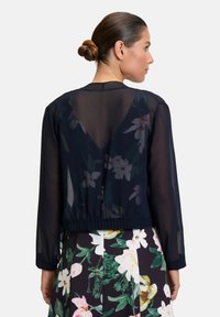 Marineblaue durchsichtige Bluse mit langen Ärmeln, elastischem Saum und einem V-förmigen Rücken. Blumendesign darunter, kombiniert mit einem bunten gedruckten Rock.