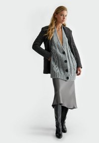 Cardigan grigio a maglia spessa con grandi bottoni neri, indossato sopra una gonna satin grigia. Il modello indossa stivali neri alti fino al ginocchio con texture.