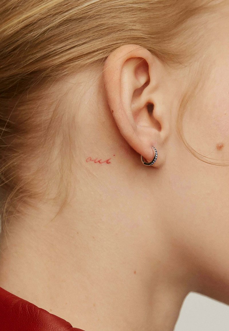 Tatuaje en el cuello que dice "oui" en tinta roja; pequeña arete de aro plateado realza la oreja. Cabello rubio claro recogido. Textura de piel suave.