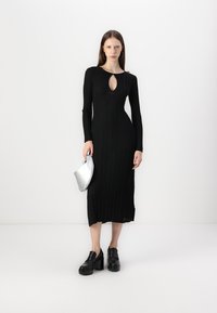 Black ribbed dress with long sleeves and a keyhole neckline. Model holds a silver bag and wears black platform shoes. Smooth texture.