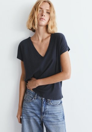 Jeune personne aux cheveux blonds mi-longs portant un haut noir à manches courtes avec un décolleté en V et un jean en denim bleu clair sur fond blanc.