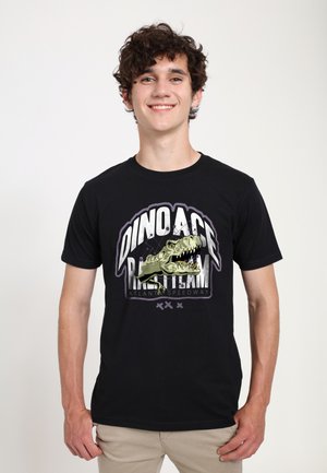 Jeune homme souriant, portant un t-shirt noir avec un dinosaure et le texte « DINO ACE RACE TEAM Atlanta Speedway » en lettres grasses.
