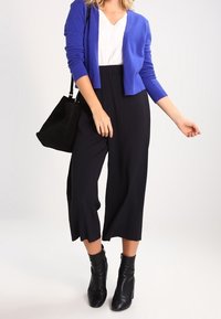 Blå kofta över en vit topp, parat med svarta capribyxor. Svarta ankelskor och en svart tote-väska. Slät tyg och avslappnad passform.