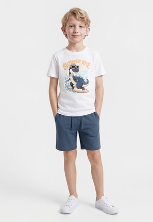 Dječak u bijeloj majici s printom dinosaurusa na dasci za surfanje i natpisom "SUMMERTIME", plavim kratkim hlačama i bijelim tenisicama, stoji s rukama u džepovima.