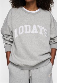 Grauer Sweatshirt mit weißer "10DAYS" Beschriftung, aus weichem Material, mit Rundhalsausschnitt und lockeren Ärmeln, kombiniert mit grauen Jogginghosen.