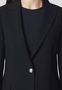 Czarna marynarka z ostrymi klapami, zapinana na pojedynczy złoty guzik, o czystych liniach. Posiada kieszeń po lewej stronie. Teksturowana tkanina.