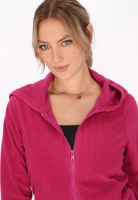 Fuksia vetoketjuhuppari, joka on valmistettu pehmeästä fleece-materiaalista, sisältää hupun ja kontrastivärisen vetoketjun sekä kapean leikkauksen. Mallilla on musta paita päällä.