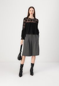 Blusa nera in pizzo con maniche lunghe e svasate sopra pantaloni corti grigi su misura. La modella indossa stivali neri alla caviglia e tiene una piccola borsa nera.