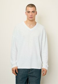 Balta ilgomis rankovėmis V formos iškirptė marškinėliai, pagaminti iš lengvos medžiagos, su laisvu siluetu ir nuleistais pečiais, derinami su tamsiomis džinsinėmis kelnėmis.