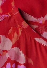 Tessuto rosso con un motivo floreale nei toni del rosa e dell'arancio. Presenta un collo arrotondato e una texture liscia, con dettagli di cucitura visibili.