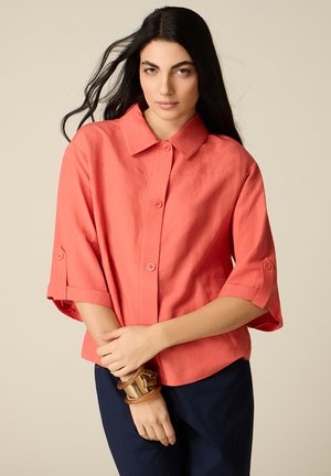 Kobieta z długimi ciemnymi włosami, ubrana w koralową koszulę zapinaną na guziki i ciemne spodnie, trzymająca nadgarstek ozdobiony dwoma masywnymi bransoletkami.