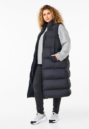 Chaleco de plumas negro largo con cuello alto, diseño acolchado, bolsillos laterales, combinado con un suéter gris y jeans oscuros, junto con zapatillas deportivas.