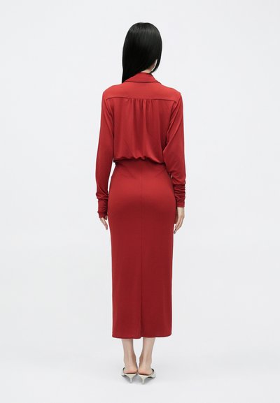 Hosszú, piros ruha sima anyagból, galléros nyakkivágással, begyűrt hátrésszel, formázott derékrésszel és egyenesen lelógó szoknyával, amely a borjúközépig ér.