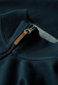 Dunkelblaues Sweatshirt aus glänzendem Material mit strukturiertem Finish, ausgestattet mit einem Reißverschluss-Kragen und einem ledernen Ziehband, das mit "Superdry" geprägt ist.