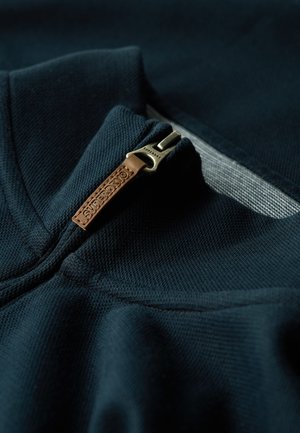 Sudadera de un oscuro azul marino con un acabado texturizado, que cuenta con un cuello con cremallera y un tirador de piel grabado con "Superdry."