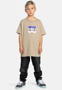 Beige t-shirt med kort ärm som har en grafisk logotyp, tillsammans med mörka jeans och grå sneakers med vita snören och teal-accenter.
