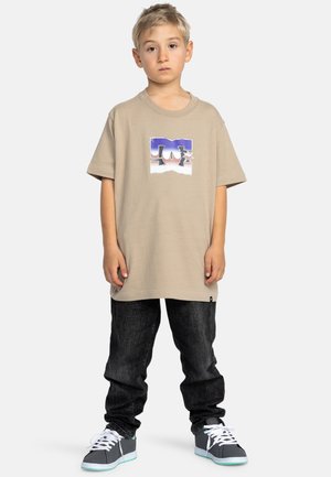 Beige t-shirt met korte mouwen met een grafisch logo, gecombineerd met donkere spijkerbroek en grijze sneakers met witte veters en teal accenten.