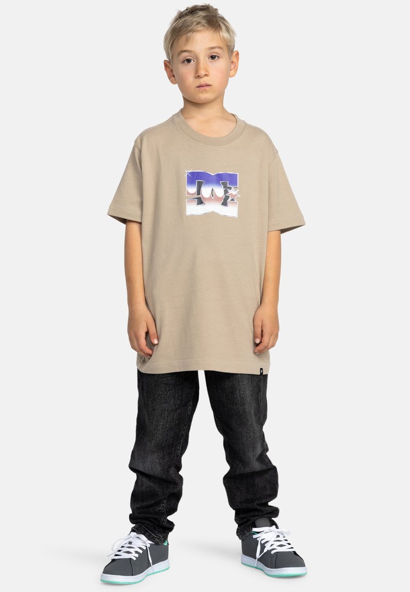 Beige t-shirt med kort ärm som har en grafisk logotyp, tillsammans med mörka jeans och grå sneakers med vita snören och teal-accenter.