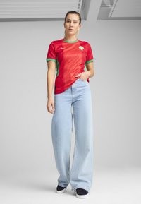 Röd och grön marockansk landslagsdräkt med en strukturerad design, kombinerad med ljusblå vida jeans och svarta sneakers.