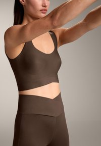 Hnedý športový crop top s širokým pásom a hlbokým výstrihom, skombinovaný s legínami s vysokým pásom, ktoré majú vrstvený dizajn a hladkú textúru.