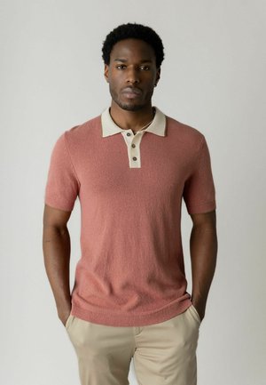 Mand med kort krøllet hår iført en pink kortærmet poloshirt med beige krave og beige bukser, stående med hænderne i lommerne.