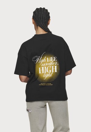 Persoon met gevlochten haar staat met de rug naar ons toe, draagt een zwart T-shirt met een gele cirkel en witte tekst met de woorden "High Life under High light."