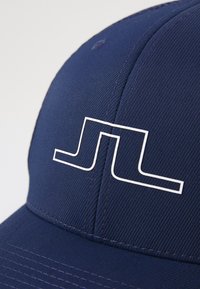 Tumed sinine müts on valmistatud tekstuursetest kangast ja sellel on valge äärisega logo kujundus. Mütsil on kumer kuiv ja nähtavad õmblused.