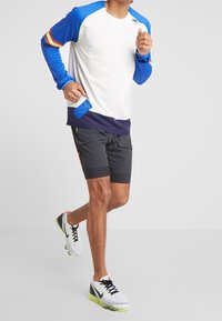 Biała sportowa koszulka z długim rękawem z niebieskimi i czerwonymi akcentami, zestawiona z czarnymi szortami. Buty są białe, z czarnymi i neonowozielonymi detalami.