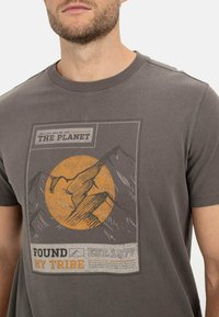 Graues Baumwoll-T-Shirt mit einem Grafikdesign von Bergen, einer orangefarbenen Sonne und dem Text "Found My Tribe EST. 1977" in kontrastierenden Farben. Kurze Ärmel.
