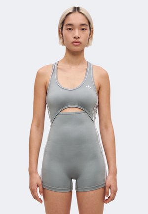 Mannequin aux cheveux blonds courts, debout face à l’avant, portant une combinaison sans manches grise Adidas avec une découpe à la taille, sur un fond uni.