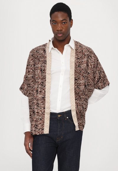 Brun mønstret kimono-stil jakke med hvit broderi langs kantene, båret over en hvit skjorte og mørke jeans. Løs passform, lett materiale.