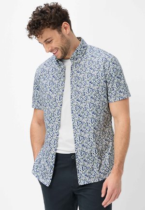 Mann lächelt, schaut nach unten, trägt ein kurzärmeliges Hemd mit blau-grünem Blumenmuster über einem weißen T-Shirt.