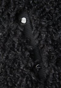 Cardigan nero con trama testurizzata che presenta una parte in tessuto soffice e una sezione lavorata a maglia liscia, chiuso con bottoni metallici lucidi. Dettagli di cuciture visibili.