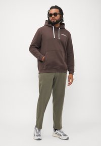 Hombre con gafas de sol, vistiendo una sudadera marrón, pantalones verde olivo y zapatillas grises, de pie con una mano en el bolsillo, contra un fondo blanco.