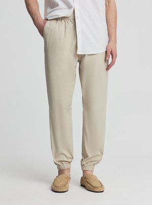 Uomo con joggers elasticizzati beige alla caviglia, mocassini slip-on color cuoio e una camicia bianca abbottonata con una mano in tasca.