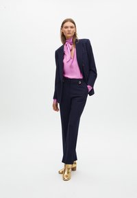 Abito navy con spalle decorate, abbinato a una blusa rosa e stivali con tacco dorati. Tessuto morbido, vestibilità su misura e bottoni distintivi.